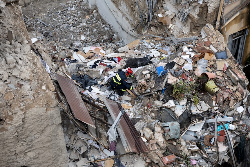 A firefighter in a red helmet and black overalls seen on top of a large pile of rubble, cluttered by concrete rocks.