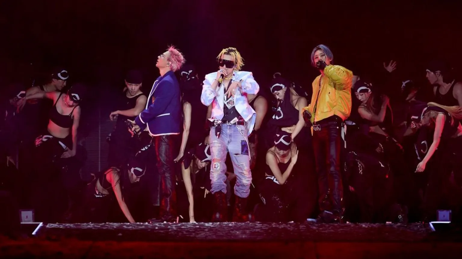 YG Entertainment Three performers stand at the centre of a stage holding handheld microphones, surrounded by a group of dancers in black outfits. One performer wears a blue jacket, another wears a white patterned jacket with sunglasses and light-coloured jeans, and the third wears a yellow jacket and dark trousers. Several dancers are positioned crouching or standing around them. The stage is lit with red and purple lighting, with a large dark screen or backdrop behind the group.