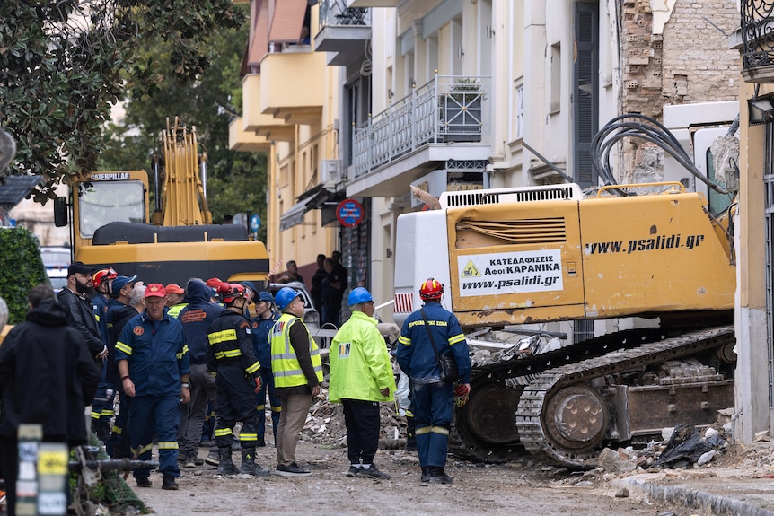 Rescue workers in blue, black and bright yellow overalls and clothing, wearing hard hats in a street next to a yellow digger.