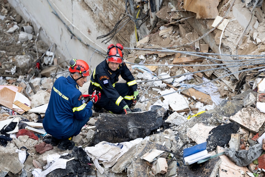 Greek firefighters in blue and black overalls crouching amid piles of rubble, broken tiles, concrete and metal bars.