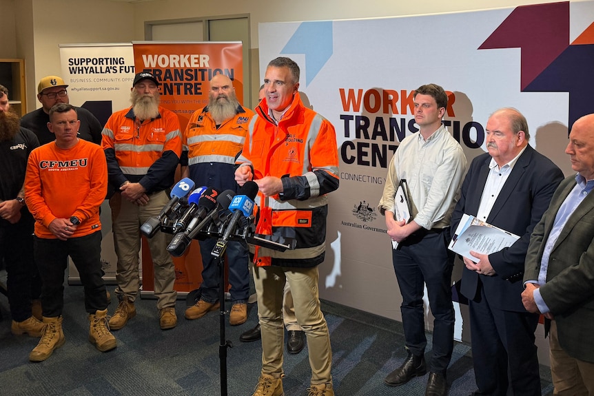 a press conference with men wearing orange hi-vis and others in suits speaking to media