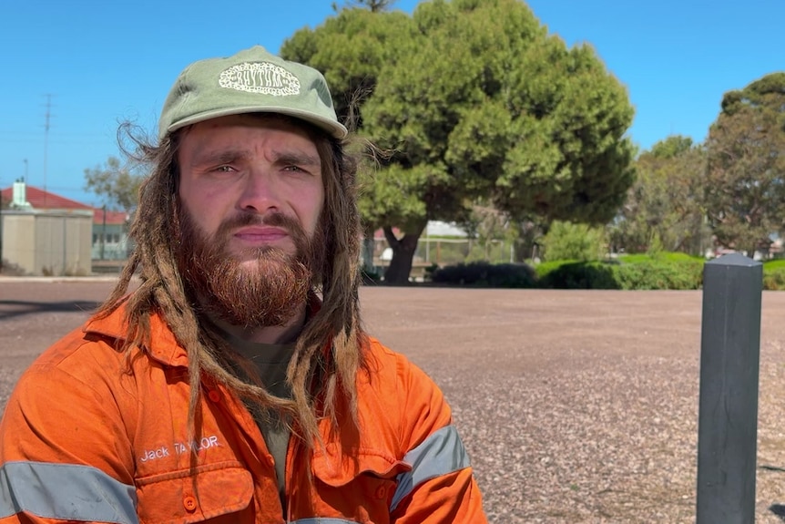 A man with dreadlocks and a beard, wearing a green cap and orange-high visibility shirt, stands in front of a vacant lot