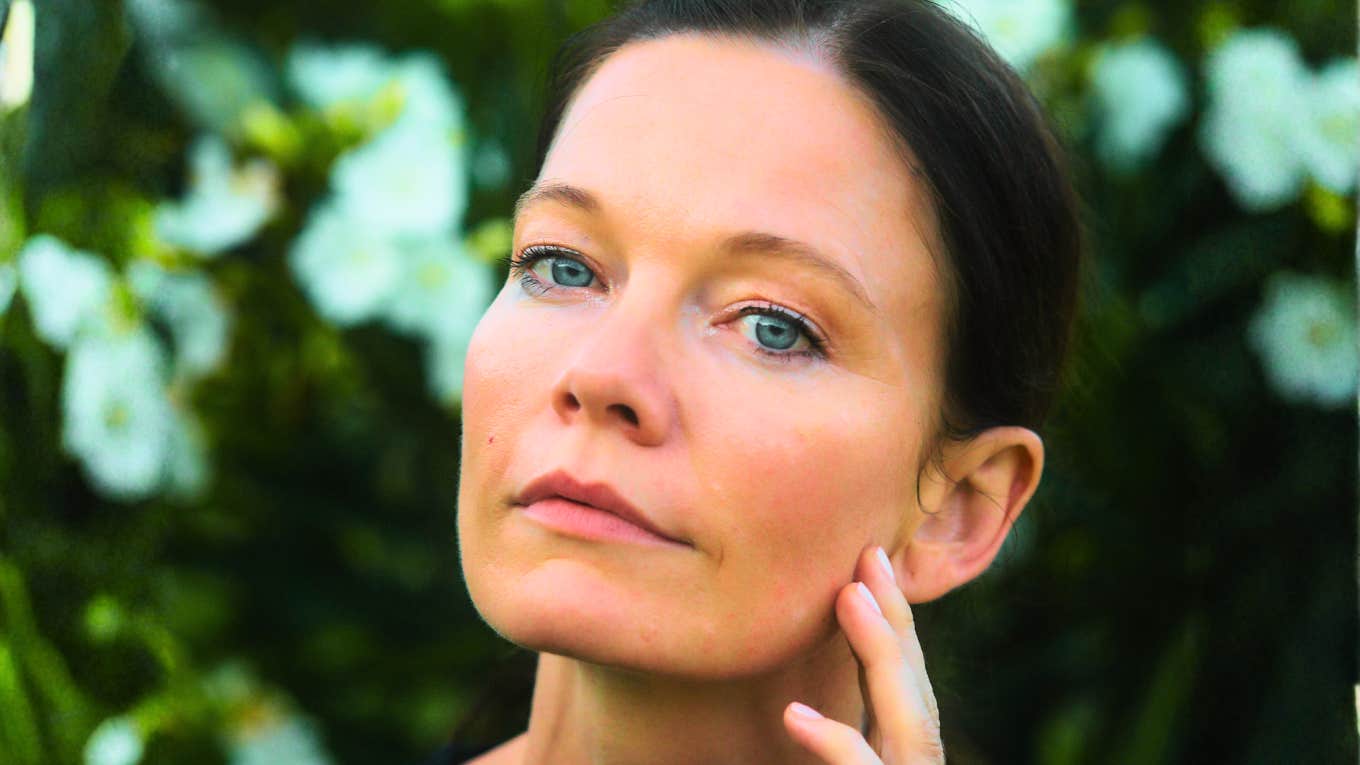A close-up portrait of a woman with light blue eyes and dark hair resting her hand on her cheek against a background of white flowers.
